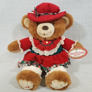 May include: A plush brown teddy bear wearing a red velvet dress with white lace trim and a matching hat. The bear has a green bodice and white stockings with a Christmas print. A tag reads "Teddy Bear Lane Christmas."