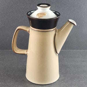 以下が含まれることがあります： 黒縁と白蓋が付いたベージュのセラミック製コーヒーポット。蓋には「Denby Fine Stoneware」と書かれた茶色のラベルが付いています。