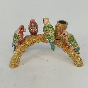 Op de afbeelding: Een goudkleurig metalen beeldje met vier kleine vogels op een gebogen tak. De vogels zijn versierd met groene, rode en doorzichtige strass-steentjes. Aan één uiteinde van de tak bevindt zich een kleine, holle cilindrische opening.