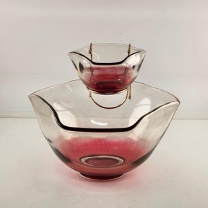May include: A vintage two-piece glass serving set. The larger bowl is clear with a pink base and rim, and the smaller bowl is suspended above it by a gold-colored metal stand. The smaller bowl is also pink. The set is designed for serving dips and snacks.
