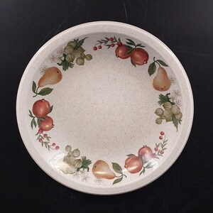 May include: A round, off-white serving plate with a speckled texture. The rim is decorated with illustrations of pears, pomegranates, gooseberries, and small white flowers. The plate has a slightly raised edge.