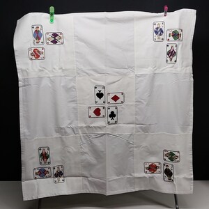 Puede incluir: Un mantel de tela blanca con diseños de cartas de juego bordados. El centro muestra cuatro ases en los palos de picas, diamantes, corazones y tréboles. Las esquinas están decoradas con varias figuras, incluyendo sotas, reinas y reyes en hilo de color.