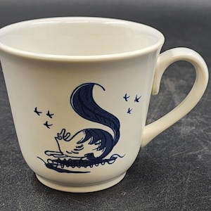 May include: A cream-colored ceramic mug featuring a dark blue graphic illustration of a stylized bird with a large, curved tail. The design includes small bird silhouettes flying above the main figure, all set against the smooth, off-white surface.