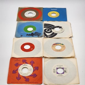 May include: A selection of vintage 45 RPM vinyl records in their original paper sleeves. The sleeves are in various colours, including blue, orange, and brown, with printed text and graphics. The records have colourful labels.