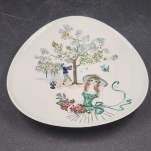May include: A white, triangular decorative plate featuring a painted scene. The scene shows a woman in a green dress and hat holding flowers, a man near a tree, and birds. The plate has a glossy finish.