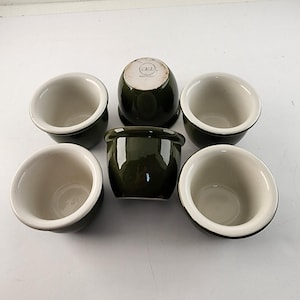 May include: Six dark green ceramic bowls with white interiors and a glossy finish. The bowls have a rounded shape with a slightly flared rim. One bowl is upside down, revealing a stamp on the bottom. The bowls are arranged on a white surface.