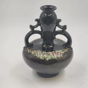 May include: A dark brown ceramic vase with a bulbous base and a tall, cylindrical neck. The vase features two curved handles and a floral design around the base. The vase is approximately 20 cm tall.