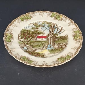 May include: A white ceramic plate featuring a rural scene with a house, trees, and a fence. The plate has a scalloped edge with a brown and green leaf pattern around the rim. The plate's diameter is approximately 25 cm.