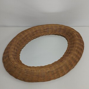 May include: Oval-shaped mirror with a woven rattan frame. The frame is a light brown color and has a textured surface. The mirror is a simple, yet elegant piece of home decor.