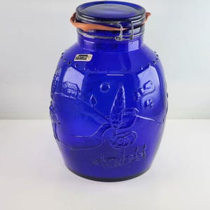 Pode incluir: Um pote de vidro azul cobalto com tampa articulada e uma alça de couro marrom. O pote tem um desenho em relevo de uma casa e flores.