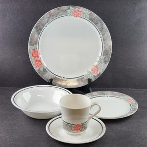 Könnte beinhalten: Ein weißes Geschirrset mit einem großen Teller, einer Schüssel, einer Untertasse, einer Tasse und einem kleineren Teller. Jedes Teil hat einen schwarzen Rand und einen dekorativen Rand mit rosa Rosen und grauen Akzenten.