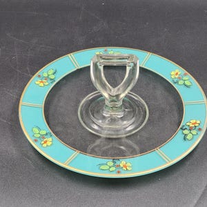 Op de afbeelding: Een heldere glazen serveerschaal met een turquoise rand. De rand is versierd met gouden strepen en bloemmotieven in geel, groen en rood. De schaal heeft een heldere glazen handgreep.