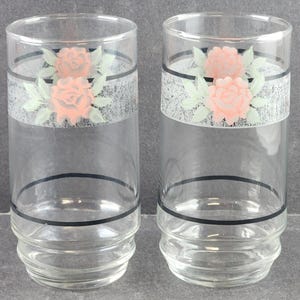 Könnte beinhalten: Zwei transparente Trinkgläser mit einem dekorativen Band, das rosafarbene Rosen und grüne Blätter zeigt. Das Band hat einen strukturierten grauen Hintergrund und wird von schwarzen Linien umrandet. Die Gläser haben ein klassisches Design.
