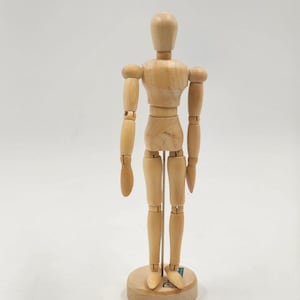 May include: A light-colored wooden articulated mannequin figure standing upright on a circular base. The figure features a smooth, natural wood grain finish with visible joints at the shoulders, elbows, wrists, hips, knees, and ankles for posing.