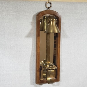May include: A vintage wall-mounted mirror with a wooden frame and a brass candle holder. The mirror is rectangular with an arched top. The candle holder has a conical shade and a decorative base. A brass ring is at the top for hanging.