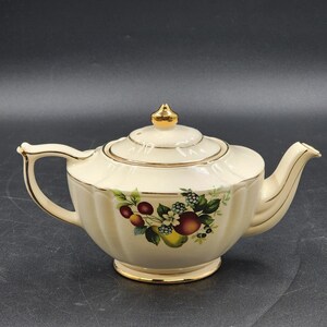 May include: A cream-colored ceramic teapot with a fluted body and a gold-trimmed lid, handle, and spout. The center features a colorful botanical illustration of various fruits, including cherries, blackberries, and a pear, with green leaves.