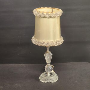 Puede incluir: Una lámpara de mesa vintage con una pantalla de color crema y una base de cristal. La pantalla tiene un borde con volantes. La base de la lámpara es de cristal transparente facetado y tiene un vástago de metal plateado. La lámpara mide unos 36 cm de altura.
