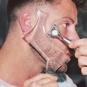 May include: A clear plastic beard shaping tool is held against a person's face while shaving with a silver razor. The tool has curved edges and a comb-like design, used to shape the beard.