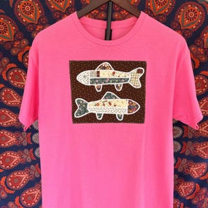 May include: A bright pink short-sleeved t-shirt featuring a rectangular patch on the chest. The patch contains two fish shapes made from various floral and plaid fabric scraps, set against a dark brown background with visible stitching.