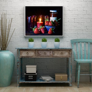 Könnte beinhalten: Ein rustikaler Holz-Konsolentisch mit einer abgewetzten weißen Oberfläche. Der Tisch hat zwei Schubladen und ein unteres Regal. Eine blaue Vase mit einem Blumenarrangement, eine weiße Box und eine kleine Holzkiste stehen auf dem Tisch. Ein blauer Stuhl steht rechts neben dem Tisch. Ein gerahmtes Bild mit einem Stillleben mit Erdbeeren, einer Kerze und einem Glas Marmelade hängt an der Wand über dem Tisch.