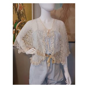 May include: A cream-colored lace capelet with a tie closure and a matching belt. The capelet is layered over a light blue outfit. The mannequin is displayed in front of a painting.