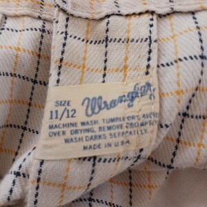 K&ouml;nnte beinhalten: Ein wei&szlig;es und gelbes kariertes Wrangler-Hemd-Etikett mit blauen N&auml;hten. Das Etikett tr&auml;gt die Gr&ouml;&szlig;e 11/12 und Anweisungen zum Waschen und Trocknen des Hemdes. Das Etikett lautet "Maschinenw&auml;sche, Trocknergeeignet, &uuml;berm&auml;&szlig;iges Trocknen vermeiden, sofort entfernen, dunkle Farben separat waschen. Hergestellt in den USA."