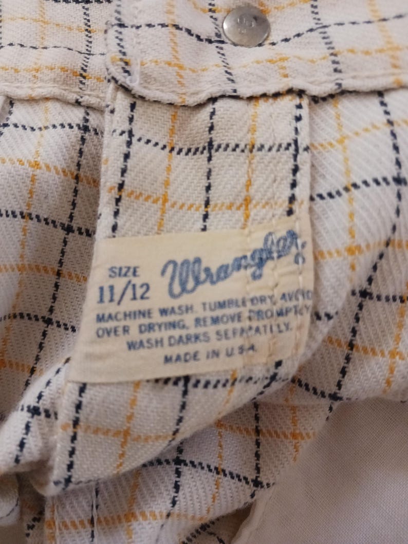 K&ouml;nnte beinhalten: Ein wei&szlig;es und gelbes kariertes Wrangler-Hemd-Etikett mit blauen N&auml;hten. Das Etikett tr&auml;gt die Gr&ouml;&szlig;e 11/12 und Anweisungen zum Waschen und Trocknen des Hemdes. Das Etikett lautet "Maschinenw&auml;sche, Trocknergeeignet, &uuml;berm&auml;&szlig;iges Trocknen vermeiden, sofort entfernen, dunkle Farben separat waschen. Hergestellt in den USA."
