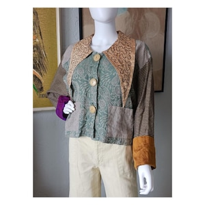 May include: A patchwork jacket with a unique design. The jacket features a paisley print, plaid, and striped patterns in shades of green, brown, and tan. It has large wooden buttons and a decorative collar. The sleeves have brown and purple accents.