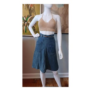 May include: A tan crochet halter top paired with a denim skirt. The skirt has a high waist, pockets, and a brown belt. The outfit is displayed on a mannequin. The background includes framed artwork.