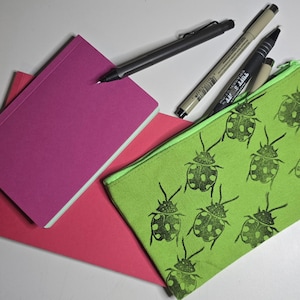 May include: A still-life composition featuring a magenta notebook, a red notepad, and a green zippered pouch with a black bug pattern. Several pens and markers are scattered around the stationery items, creating a colorful and organized workspace.
