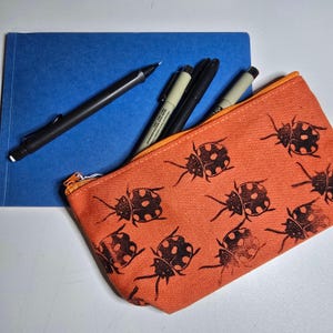 Può includere: Un astuccio arancione con un motivo di coccinelle nere, un quaderno blu e diverse penne. L'astuccio ha una cerniera ed è pieno di penne di vari colori e dimensioni. Il quaderno è di colore blu unito.