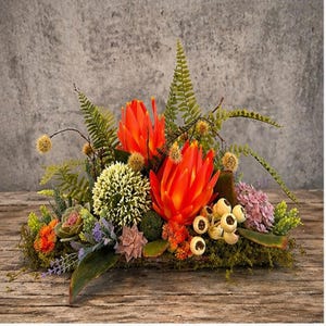 May include: A vibrant floral arrangement featuring large orange protea flowers, green ferns, and various other colorful blooms. The arrangement is set on a wooden base, creating a natural and decorative centerpiece.