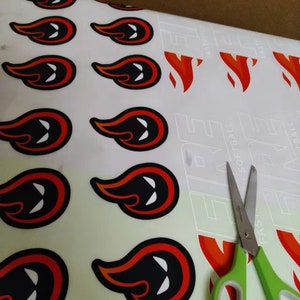 May include: A sheet of adhesive decals featuring a repeating flame graphic with a black center and white eye-like shapes, outlined in orange and black. A pair of green-handled scissors rests on the right side of the sheet, which also displays the text FIRE.