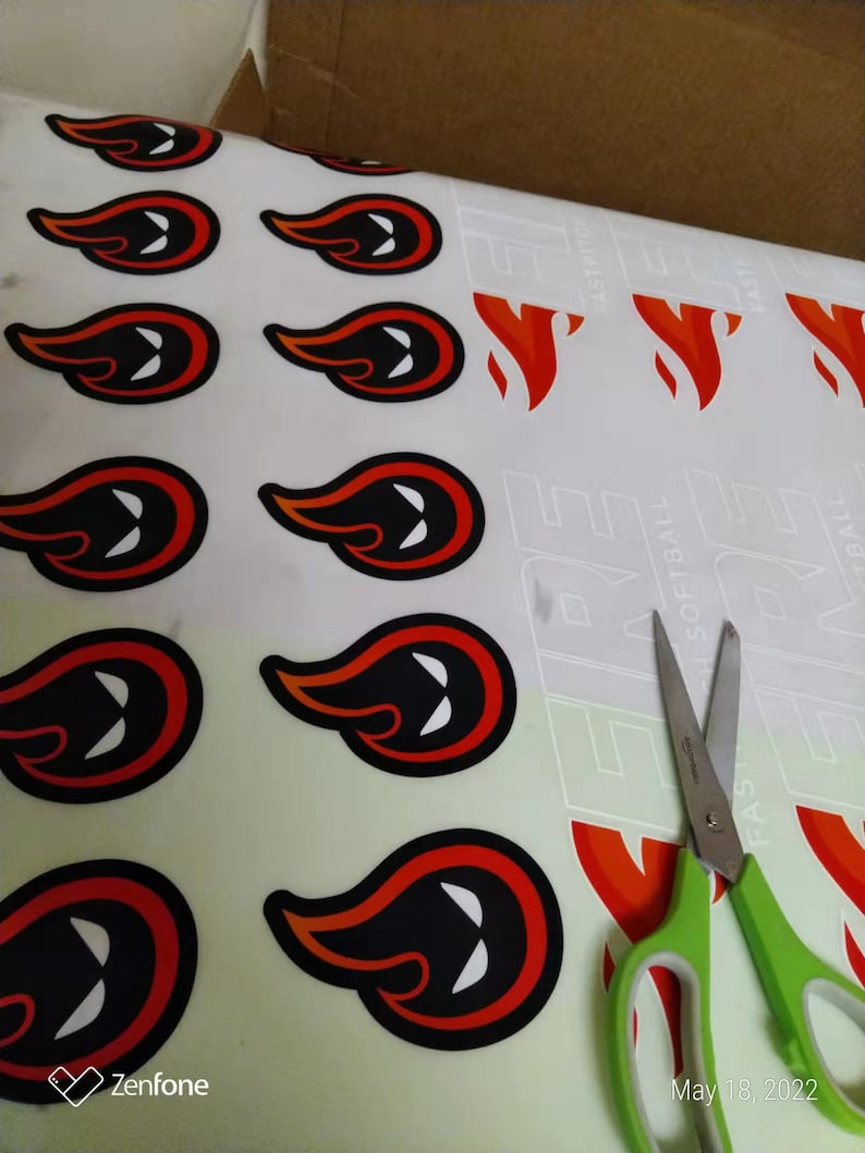 May include: A sheet of adhesive decals featuring a repeating flame graphic with a black center and white eye-like shapes, outlined in orange and black. A pair of green-handled scissors rests on the right side of the sheet, which also displays the text FIRE.