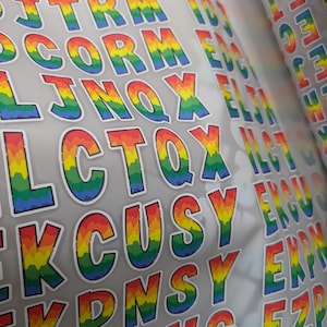 May include: A sheet of adhesive stickers featuring individual capital letters in a rainbow gradient pattern. Each letter has a white border and displays a vertical color transition from red at the top to yellow, green, and blue at the bottom.