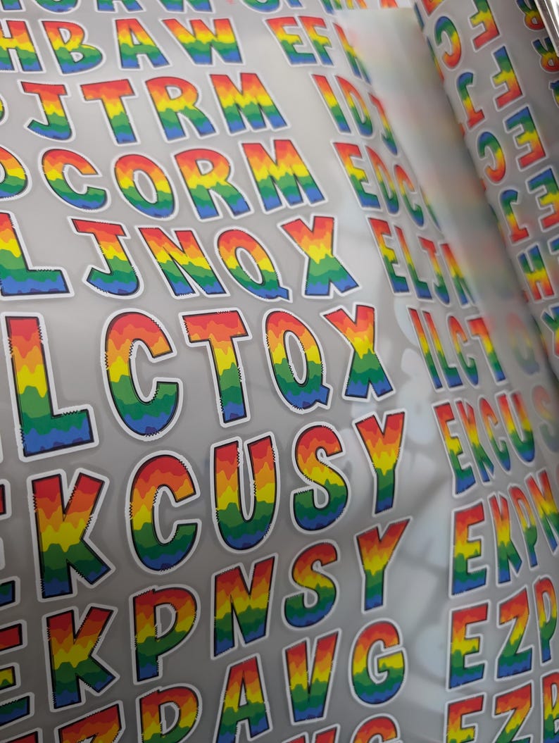May include: A sheet of adhesive stickers featuring individual capital letters in a rainbow gradient pattern. Each letter has a white border and displays a vertical color transition from red at the top to yellow, green, and blue at the bottom.