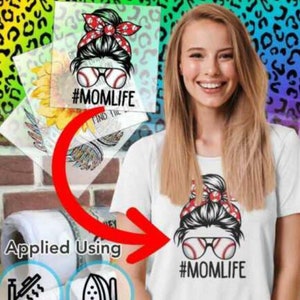 May include: A white t-shirt with a black and white graphic of a woman with a messy bun, sunglasses, and a baseball. The text "#MOMLIFE" is below the graphic. The graphic is applied to the shirt using an iron.