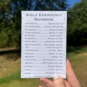 May include: A white card with the text "BIBLE EMERGENCY NUMBERS" and a list of Bible verses for various situations. The card is held against a blurred green background.