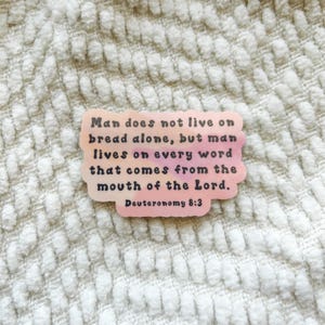 May include: A sticker with a pink and peach gradient background. The sticker has the text "Man does not live on bread alone, but man lives on every word that comes from the mouth of the Lord. Deuteronomy 8:3."
