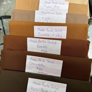 May include: A stack of fabric swatches in various shades of brown, labelled with the brand name "Moda Bella Solid" and colour names such as "Tan", "Paper Bag", "Weathered Teak", "Sienna", "Earth", "Chocolate", and "Betty's Brown". Each swatch has a corresponding number.
