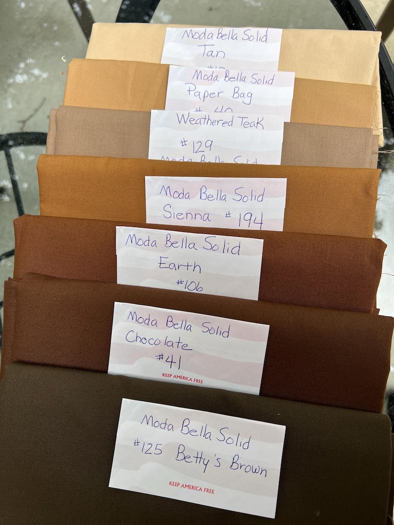 May include: A stack of fabric swatches in various shades of brown, labelled with the brand name "Moda Bella Solid" and colour names such as "Tan", "Paper Bag", "Weathered Teak", "Sienna", "Earth", "Chocolate", and "Betty's Brown". Each swatch has a corresponding number.