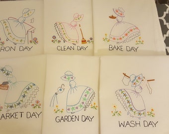 May include: Set of six embroidered tea towels featuring a woman in a dress performing different household tasks. Each towel has a different day of the week embroidered on it, such as "Iron Day", "Clean Day", "Bake Day", "Market Day", "Garden Day", and "Wash Day".