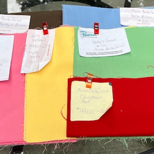 May include: An assortment of Moda Bella Solids fabric swatches in various colours, including pink, yellow, green, blue, and red. Each swatch has a handwritten label with the colour name, fabric number, and Moda Bella Solids branding.