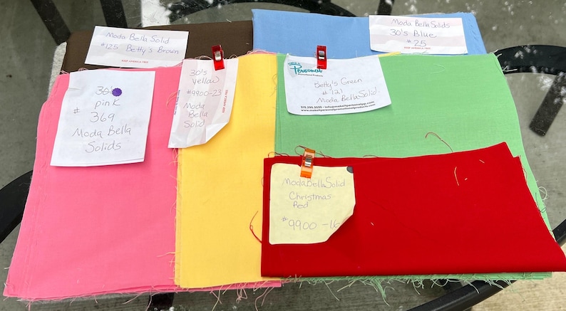 May include: An assortment of Moda Bella Solids fabric swatches in various colours, including pink, yellow, green, blue, and red. Each swatch has a handwritten label with the colour name, fabric number, and Moda Bella Solids branding.