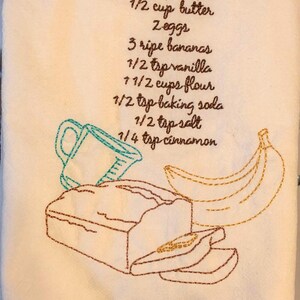 May include: A white kitchen towel with an embroidered recipe for banana bread. The recipe includes ingredients like sugar, butter, eggs, bananas, vanilla, flour, baking soda, salt, and cinnamon. The towel also features an embroidered illustration of a loaf of banana bread, bananas, and a measuring cup.
