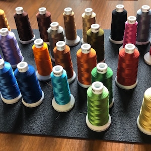 May include: A collection of spools of embroidery thread in various colours, including gold, blue, green, red, and purple. The spools are arranged on a black surface.