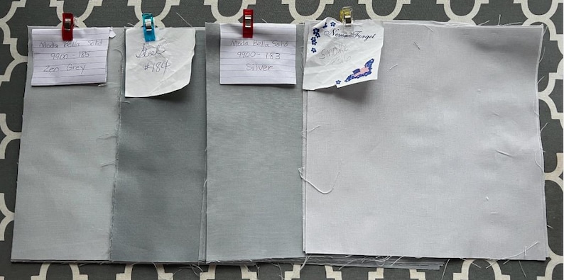 May include: Four fabric swatches in different shades of grey. The swatches are labelled with the fabric name, colour, and number. The swatches are arranged from lightest to darkest.