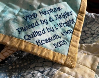 May include: A quilted fabric corner featuring a light blue and white marbled pattern with a gold-toned border. Embroidered dark blue text reads: VRD Neptune, Pieced by S. Zeigler, Quilted by J. Wright, Kossuth, Iowa, 2025, stitched onto the fabric.