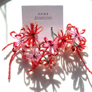 Puede incluir: Pendientes florales de cuentas rojas, rosas y transparentes, con detalles verdes. Los pendientes están sujetos a una tarjeta blanca con la marca "EDMÉ" y el texto "Bijou de perle artisanal Handcrafted bead jewelry."