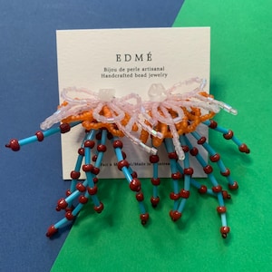 May include: A pair of beaded earrings on a white display card labeled EDME Bijou de perle artisanal Handcrafted bead jewelry. The earrings feature translucent light pink loops, orange circular beads, and dangling strands of bright blue tubes with small dark red beads.